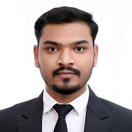 A man wearing a black suit and tie looks into the camera.